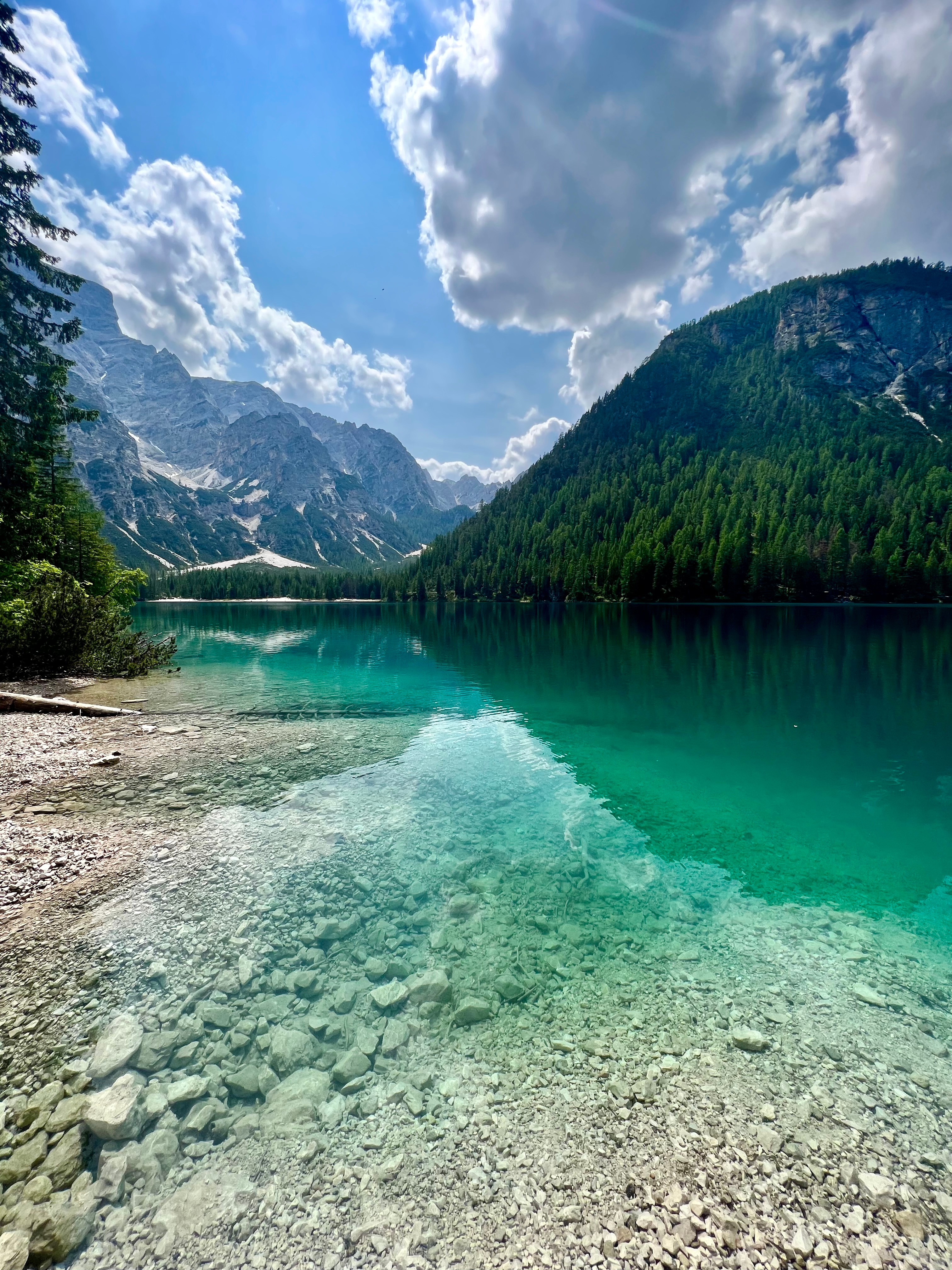 A tranquil mountain lake, framed by lush green forests and dramatic rocky peaks, reflects vibrant turquoise waters under a bright blue sky dotted with fluffy white clouds.