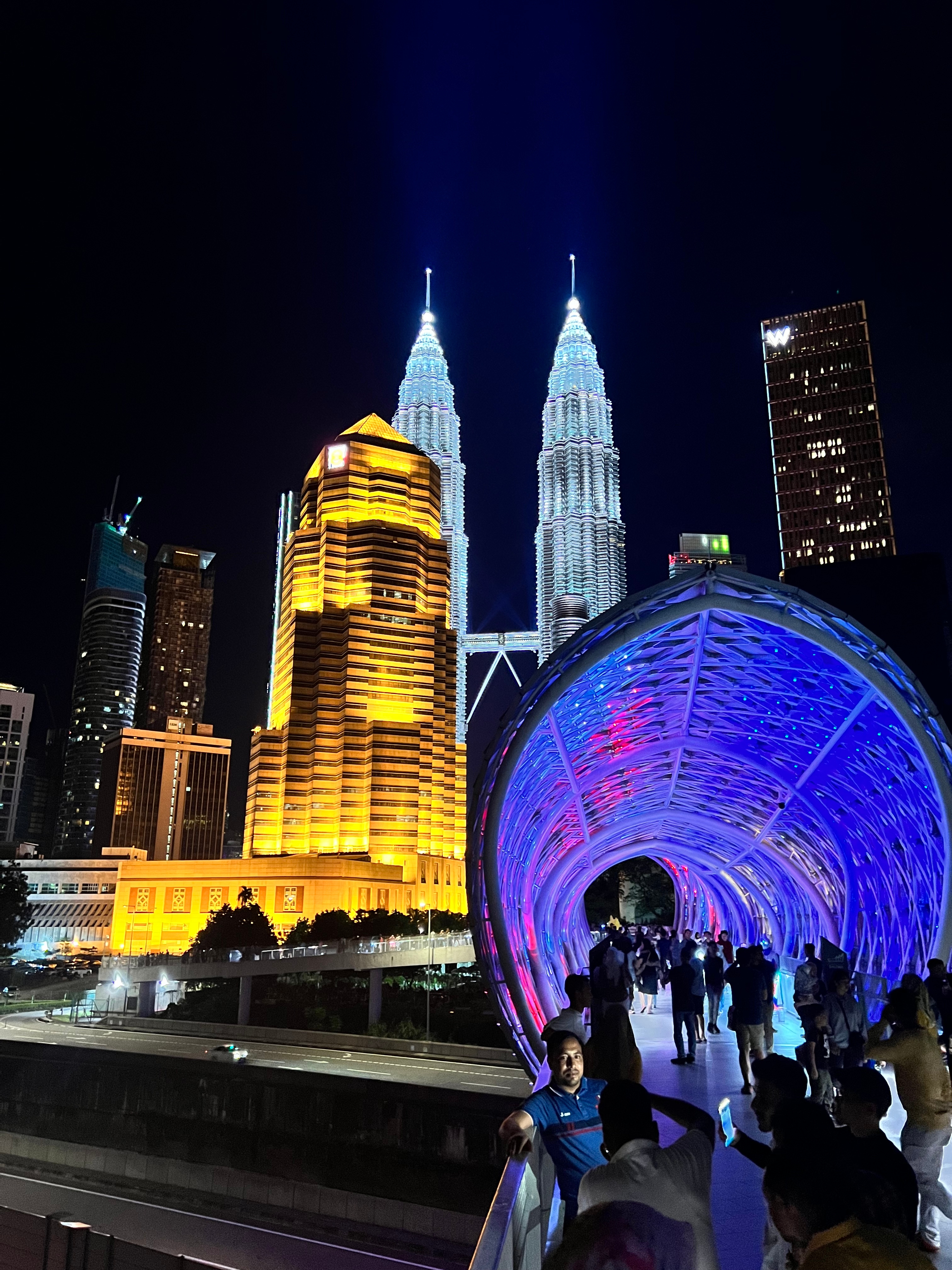 A vibrant night scene captures the iconic Petronas Towers illuminated against the dark sky, flanked by modern skyscrapers, while a bustling crowd strolls along a futuristic, colorful pedestrian bridge that glows in shades of blue and purple.