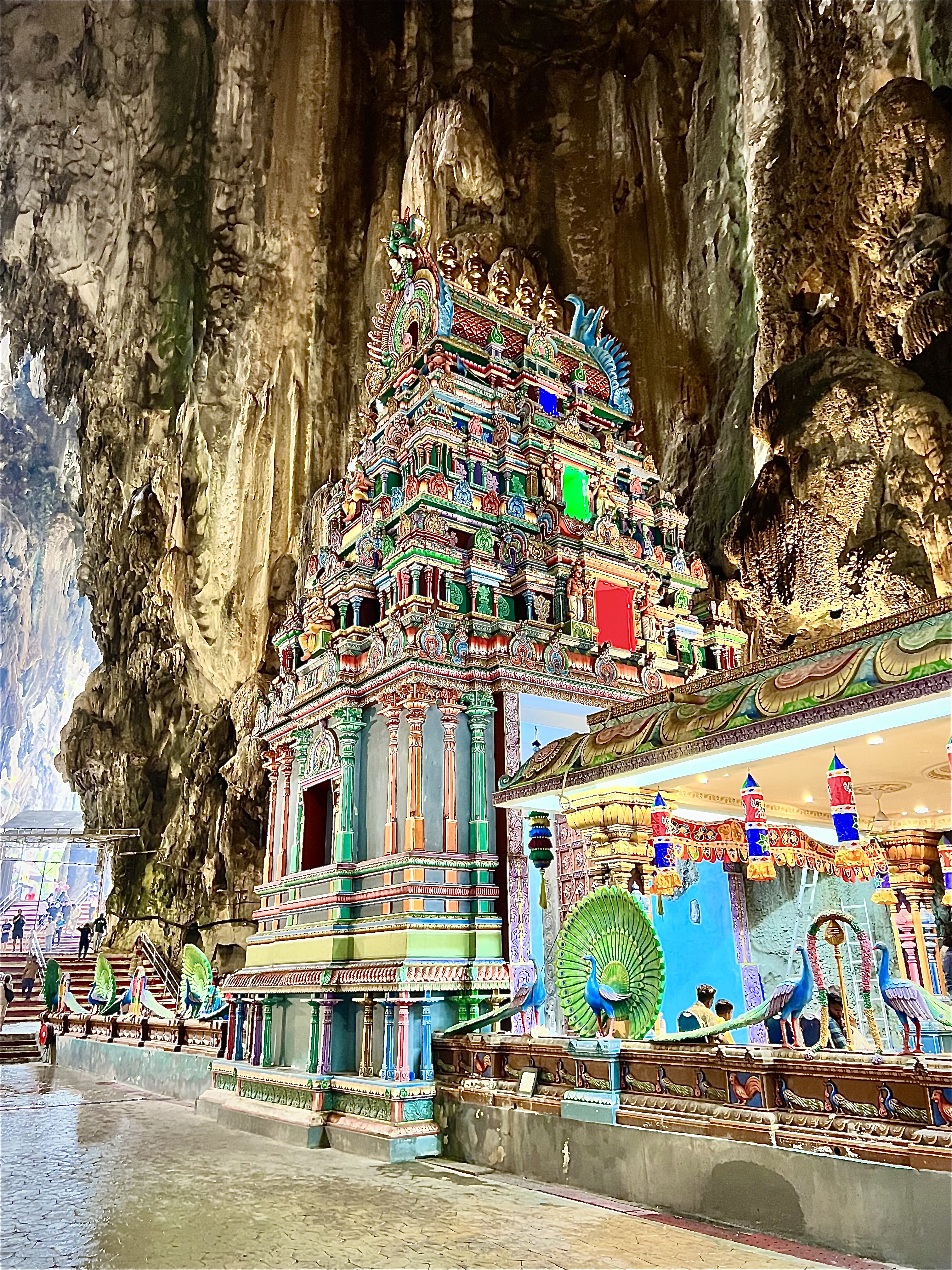The image captures an intricately designed Hindu temple adorned with vibrant colors and decorative elements, nestled within the expansive, rocky interior of Batu Caves, illuminated by soft natural light filtering through the cave's openings, while visitors explore the sacred space.