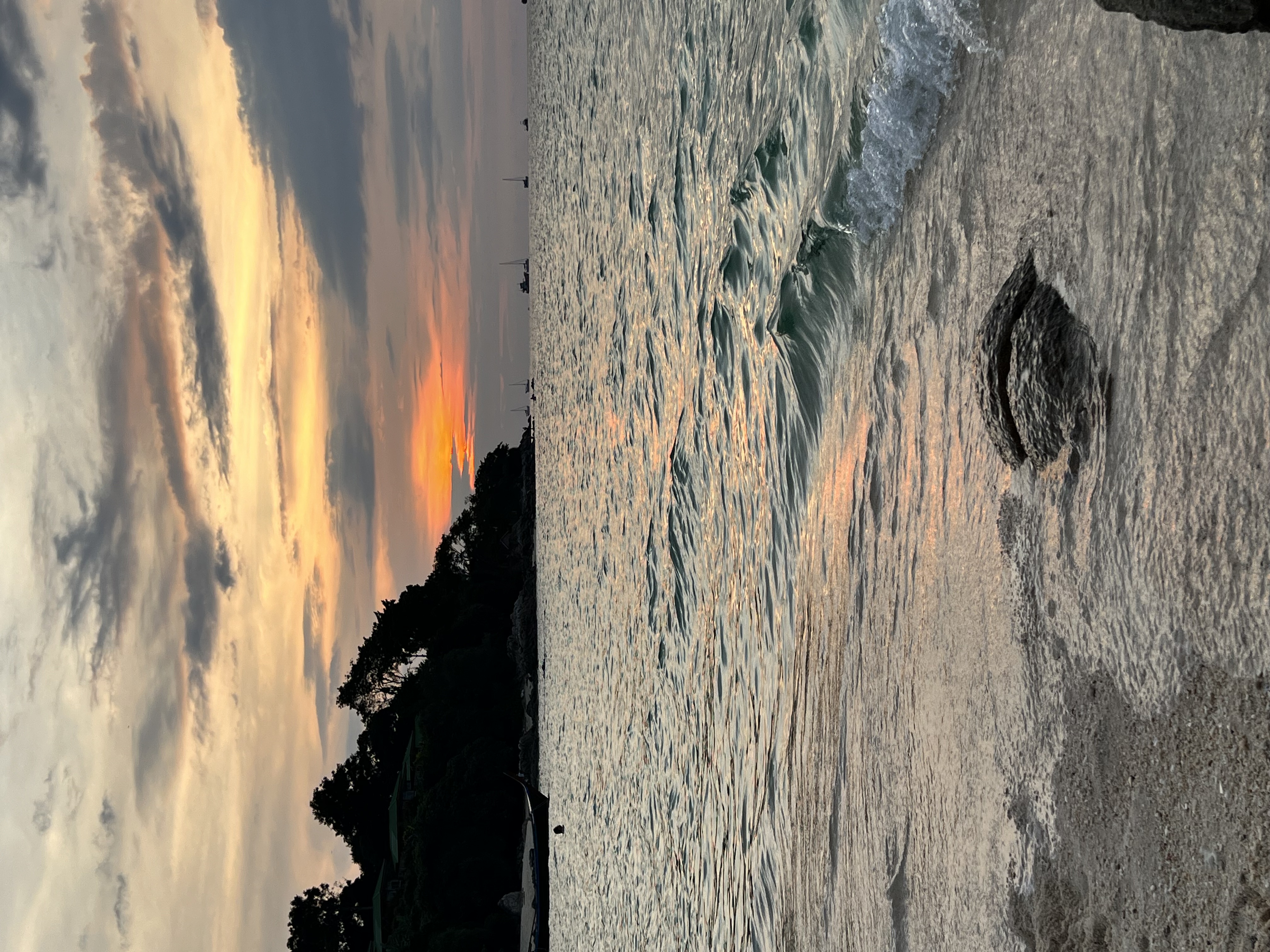 A tranquil beach scene at sunset, featuring gentle waves lapping against a sandy shore, with silhouetted trees and distant boats outlined against a vibrant sky painted in hues of orange, pink, and gray.