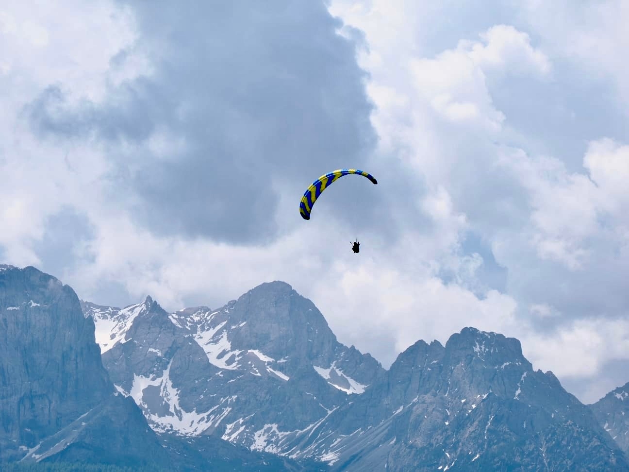 A paraglider gracefully hovers above a dramatic landscape of snow-capped mountain peaks under a sky filled with soft, billowing clouds, capturing a sense of adventure and tranquility in nature.