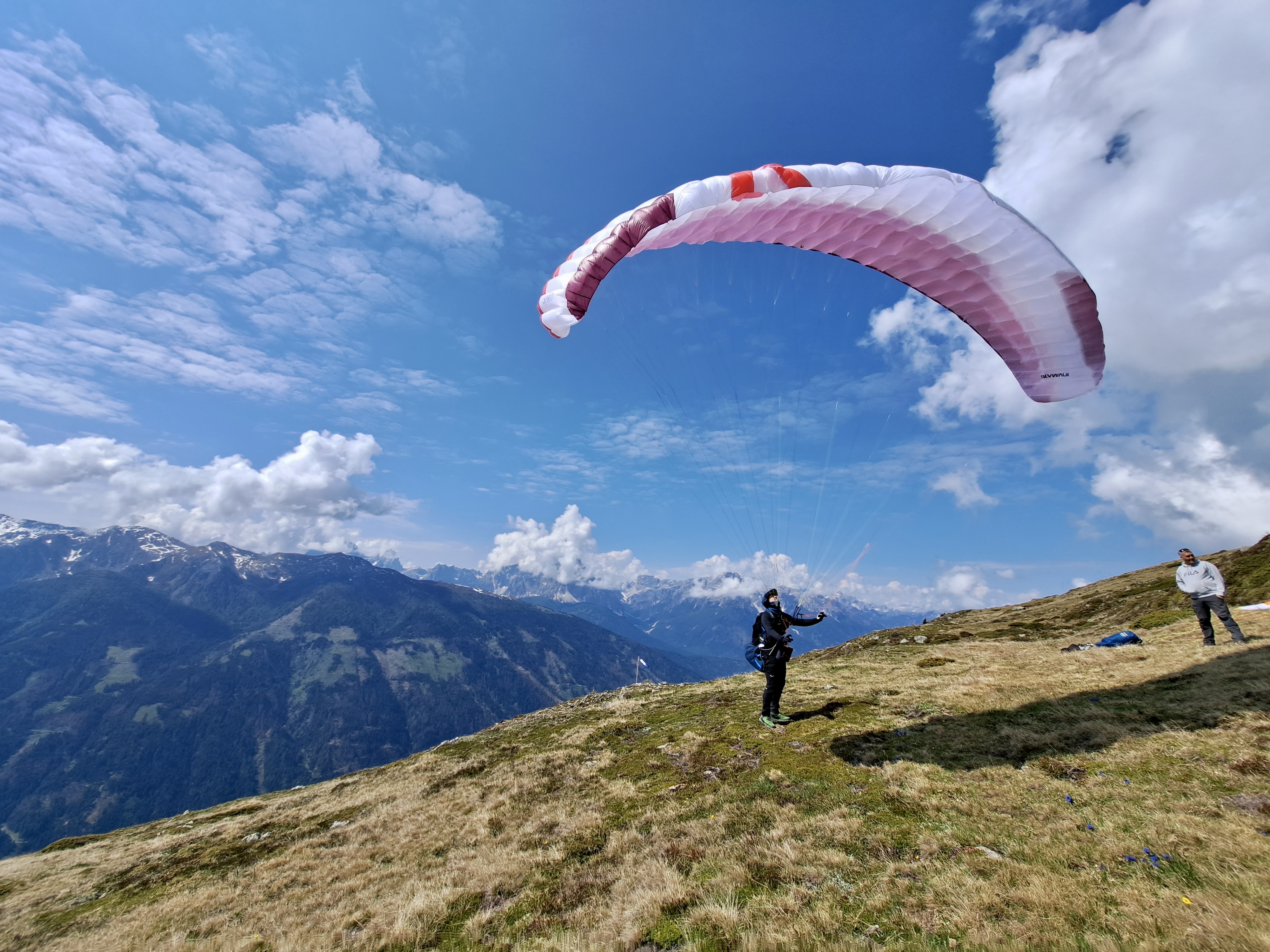 A paraglider stands on a grassy mountain slope, preparing to take off under a bright blue sky dotted with fluffy white clouds, while a companion watches nearby, with stunning snow-capped peaks in the background.